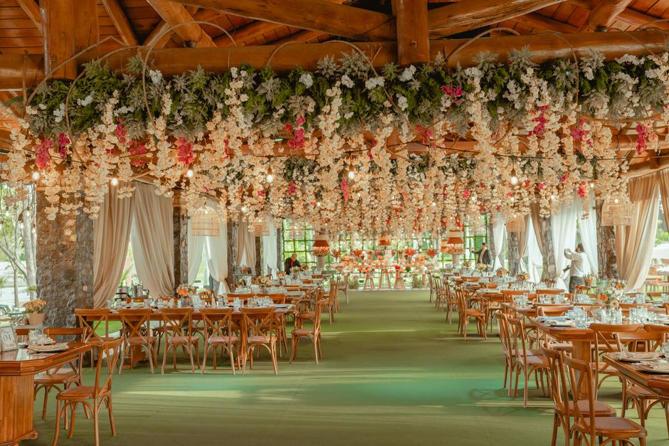 A spacious event venue decorated with an extensive floral canopy of white and pink blossoms hanging from the wooden ceiling, creating a lush, romantic atmosphere. The room features large windows drape