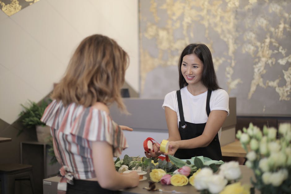 A spacious event venue decorated with an extensive floral canopy of white and pink blossoms hanging from the wooden ceiling, creating a lush, romantic atmosphere. The room features large windows drape