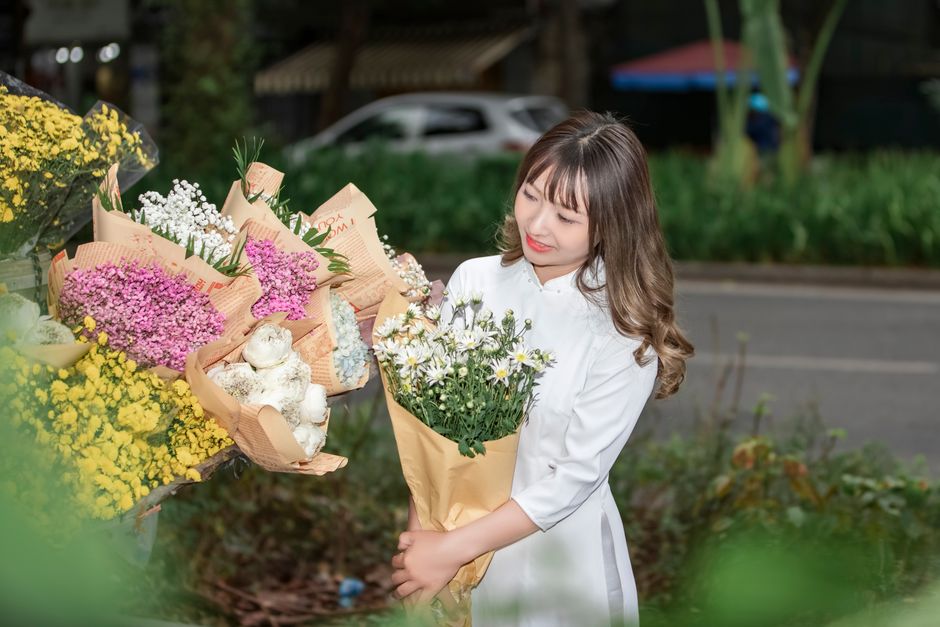 A delivery person wearing a red cap and a reflective yellow vest is holding a vibrant bouquet of white roses and green foliage in front of him, smiling at the camera. The background shows a suburban s