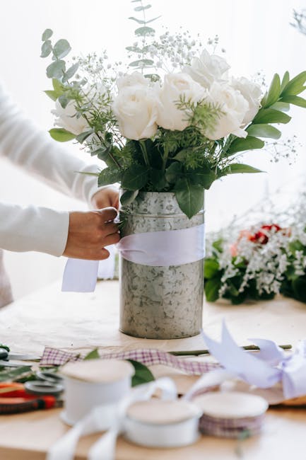 A person working with fresh white roses and green foliage in a silver vase, arranging a floral bouquet indoors under soft lighting. The roses have full, layered petals with a pristine appearance, and 