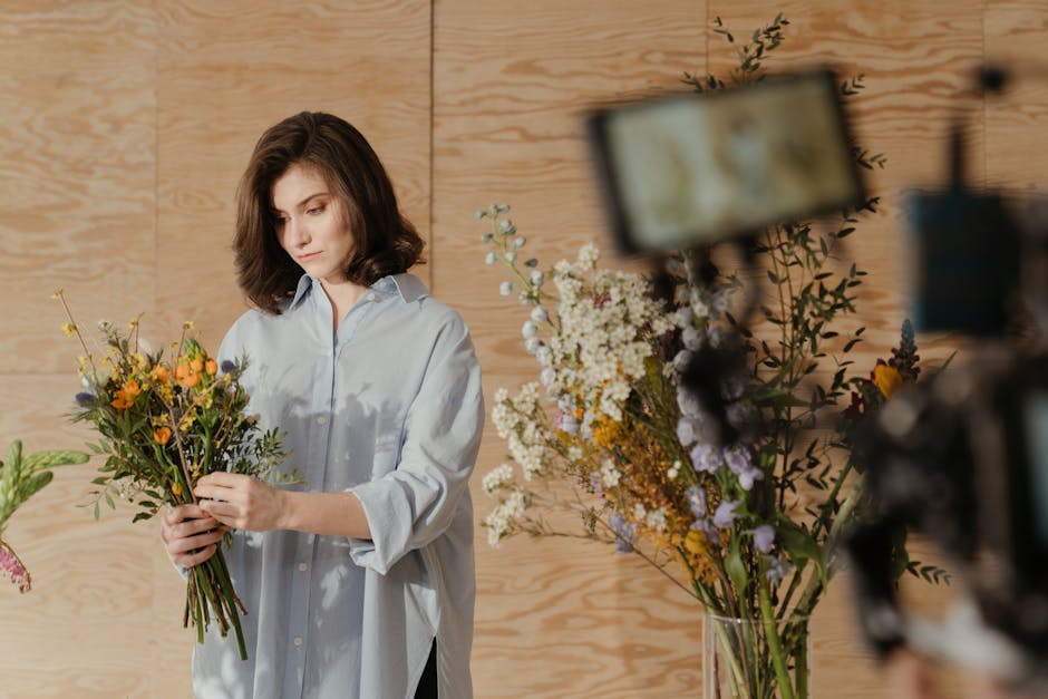 A person arranging a floral bouquet in a well-lit space, with soft pink and white peonies and green foliage spread across a wooden table. The individual, wearing a blue shirt, is carefully handling th