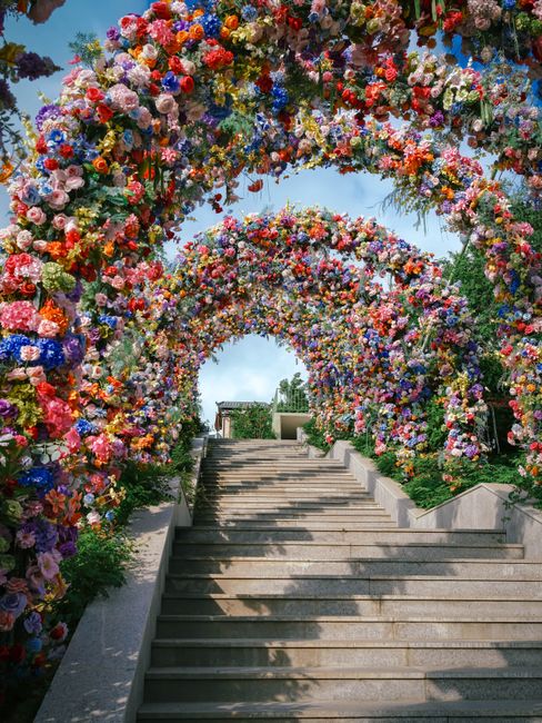 A vibrant floral display featuring an arrangement of fresh flowers with soft pink and white blossoms, including chrysanthemums and roses, cascading on a delicate, twisting wire structure. The arrangem