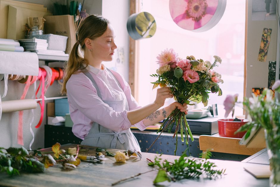 A florist from flowersallday.co.uk is in a flower shop, holding a bouquet of pale peach roses accented with green foliage. The bouquet is wrapped with a simple red floral wire, and the flowers appear 
