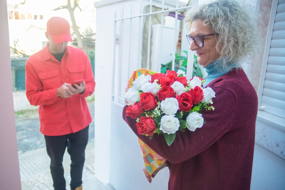 A woman with long dark hair, dressed in a white sleeveless top, is smiling while receiving a bouquet of fresh flowers wrapped in pink paper from another individual at a floral shop. The bouquet contai