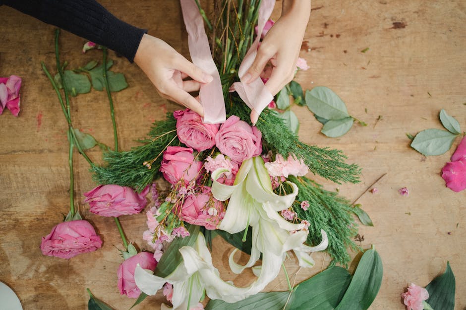 A close-up view of a meticulously crafted hand-tied bouquet featuring soft peach hydrangeas as the focal flowers, complemented by cream and white roses, and accented with sprigs of dark green foliage.