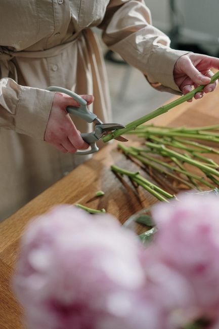 Close-up of a person’s hands arranging a bouquet of fresh flowers on a wooden floor. The bouquet includes white orchids with pink and yellow centers, pink calla lilies, and various green foliage. Th